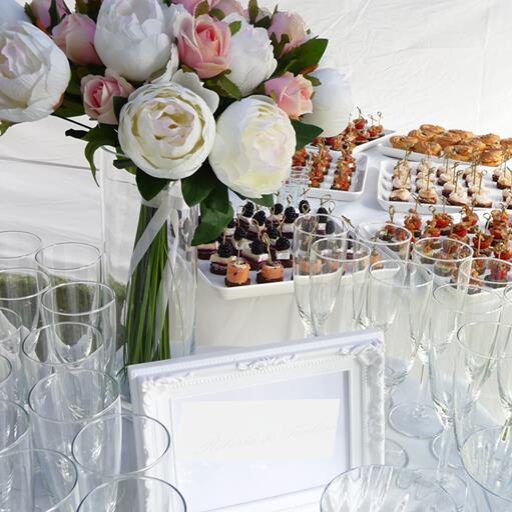 baltų staltiesių fone desertai, vaišės, šampano taurės, pastelinių gėlių puokštės- šamapno staliuko fragmentas