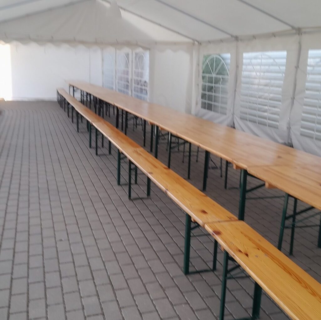 baltos palapinės ant trinkelių pastatyti mediniai stalai ir suolai, viskas yra baltoje palapinėje