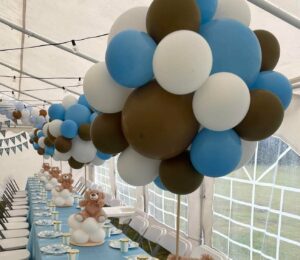 baltos palapinės viduje stovi stalas, padengtas mėlyna staltiese, ant kurios sudėtas lėkštės ir papuošimai- rudi meškiukai ir įvairiaspalviai balionai