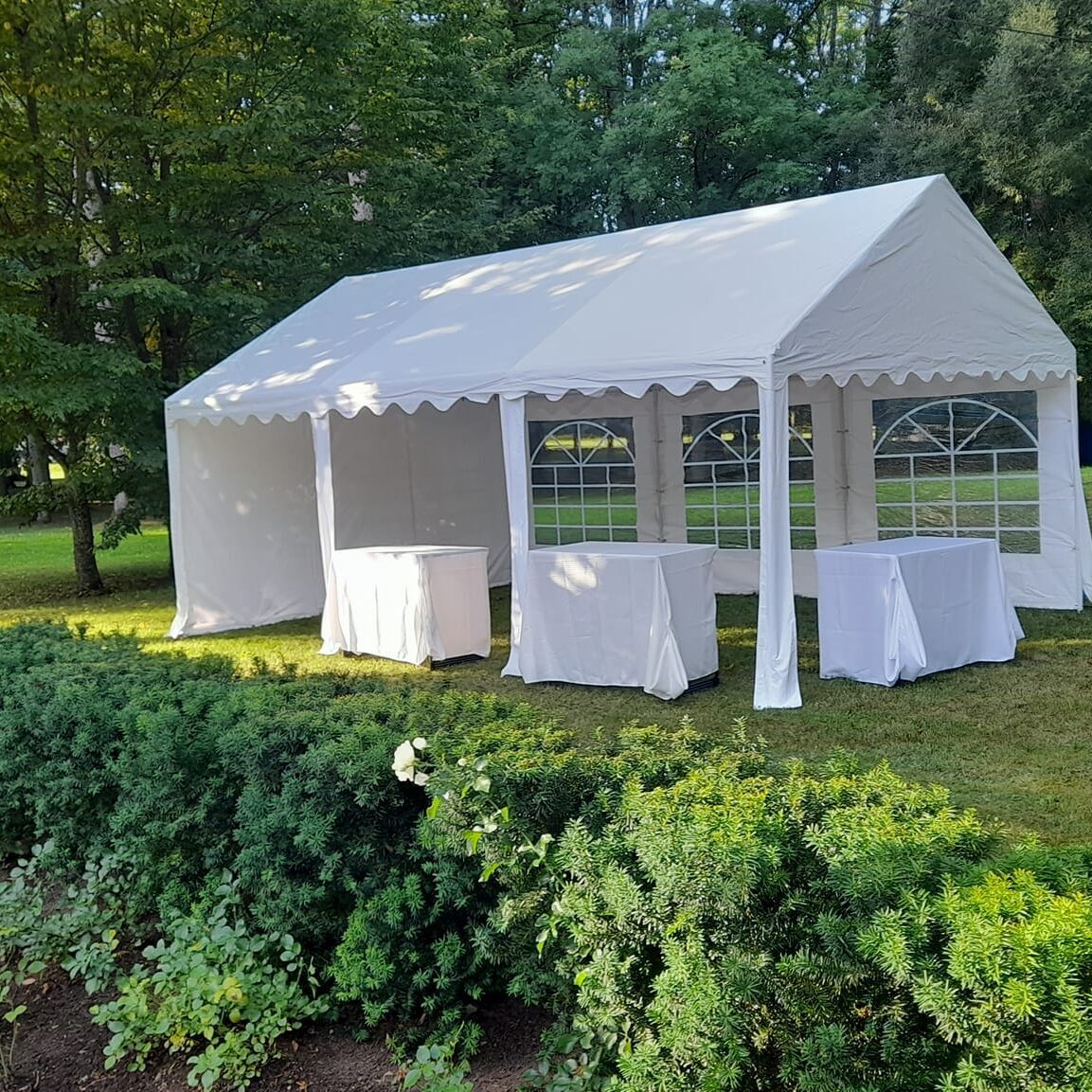 pievoje stovi balta atvira palapinė, joje stovi trys stalai, uždengti baltomis staltiesėmis