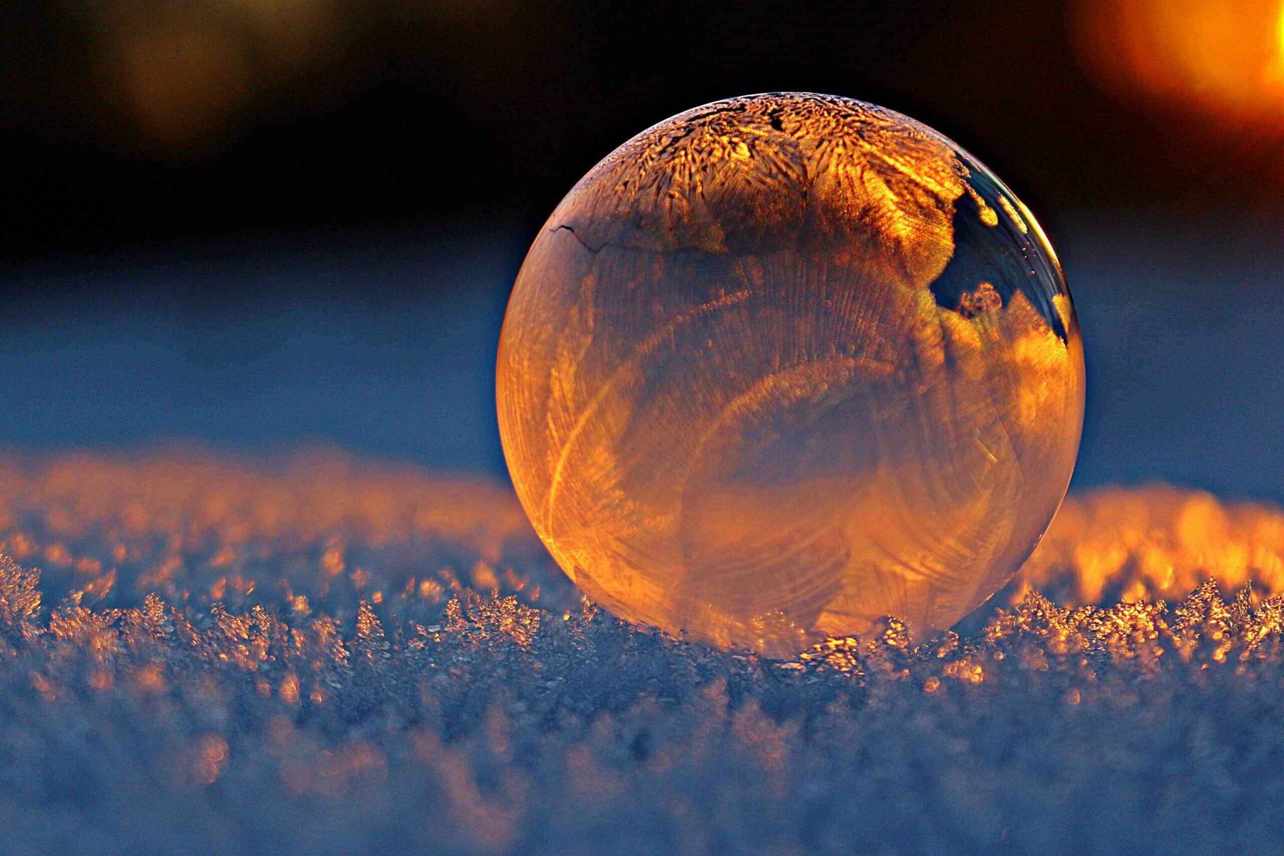 Front Page Close-up shot of a frozen bubble with warm reflections resting on a snowy surface at twilight.