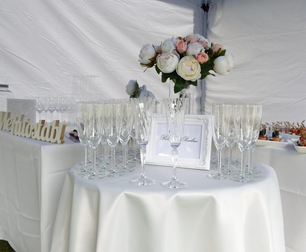 šampano staliukas palapinėje Baltos palapinės viduje baltomis staltiesėmis dengti stalai, ant kurių stovi šampano taurės, puošnūs desertai ir užkandžiai, stovi gėlių puokštės
